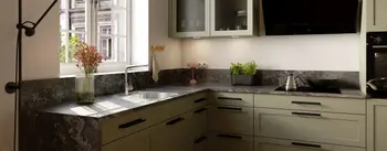 Klassische L‑Küche in Salbeigrün mit Rahmenfronten, dunkler Stein-Arbeitsplatte und großem Fenster; Doppelspüle mit Chromarmatur, Glas-Hängeschränke, schwarze Schräghaube, Induktionsfeld mit Kessel und Wok, Kräutertopf, Holzfußboden.